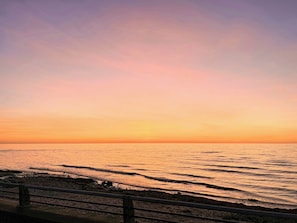 Ein Sonnenuntergang über einem ruhigen Meer mit einer felsigen Küste und einem Metallgeländer im Vordergrund.
