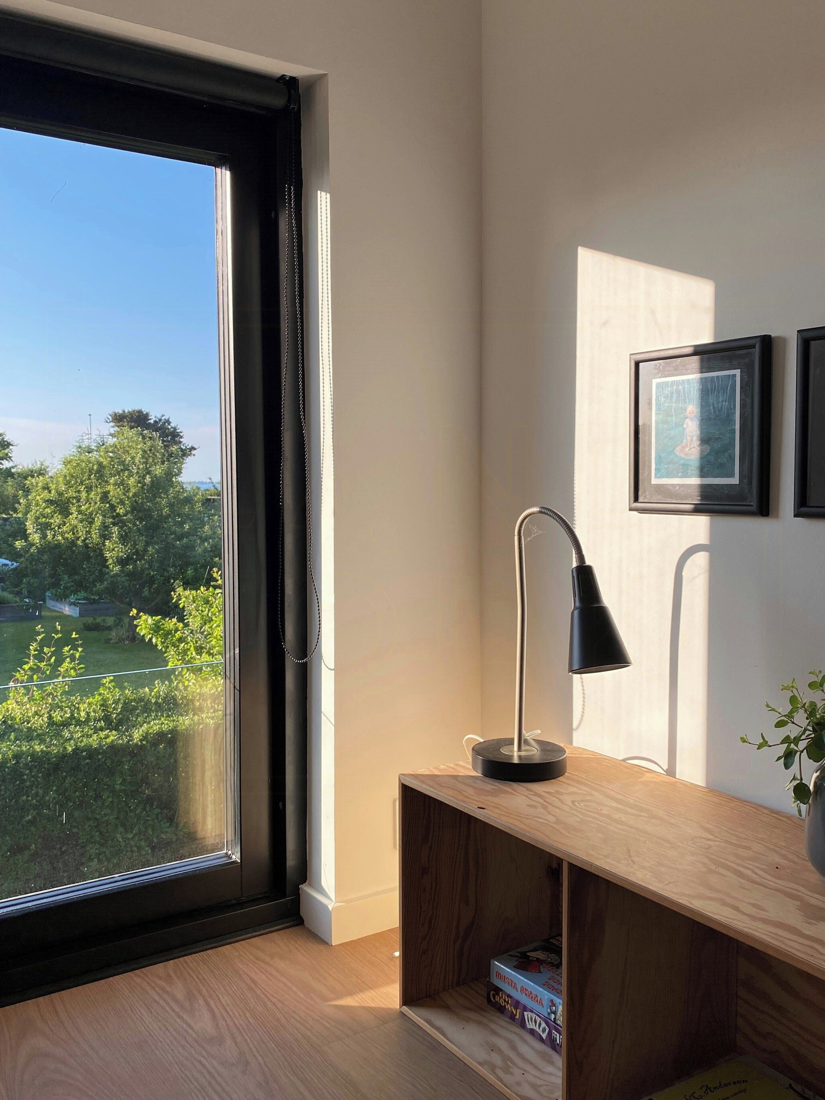 Une pièce avec une grande fenêtre, un bureau avec une lampe et des tableaux encadrés accrochés au mur.