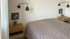 Ein Schlafzimmer mit einem Bett, zwei Nachttischen, einer Wandleuchte, einem gerahmten Bild und einer Zimmerpflanze.