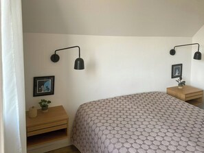 Ein Schlafzimmer mit einem Bett, zwei Nachttischen, einer Wandleuchte, einem gerahmten Bild und einer Zimmerpflanze.