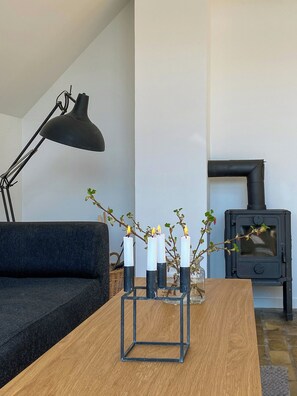 Ein Wohnzimmer mit einer Couch, einem Holztisch, einer schwarzen Stehlampe, einem schwarzen Kaminofen und einer Vase mit Kerzen.