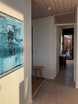 A hallway with a modern abstract painting, a wooden stool, and a view of a garden through a door.
