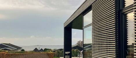 A modern house with a large glass window, a wooden deck, and a view of the sea.