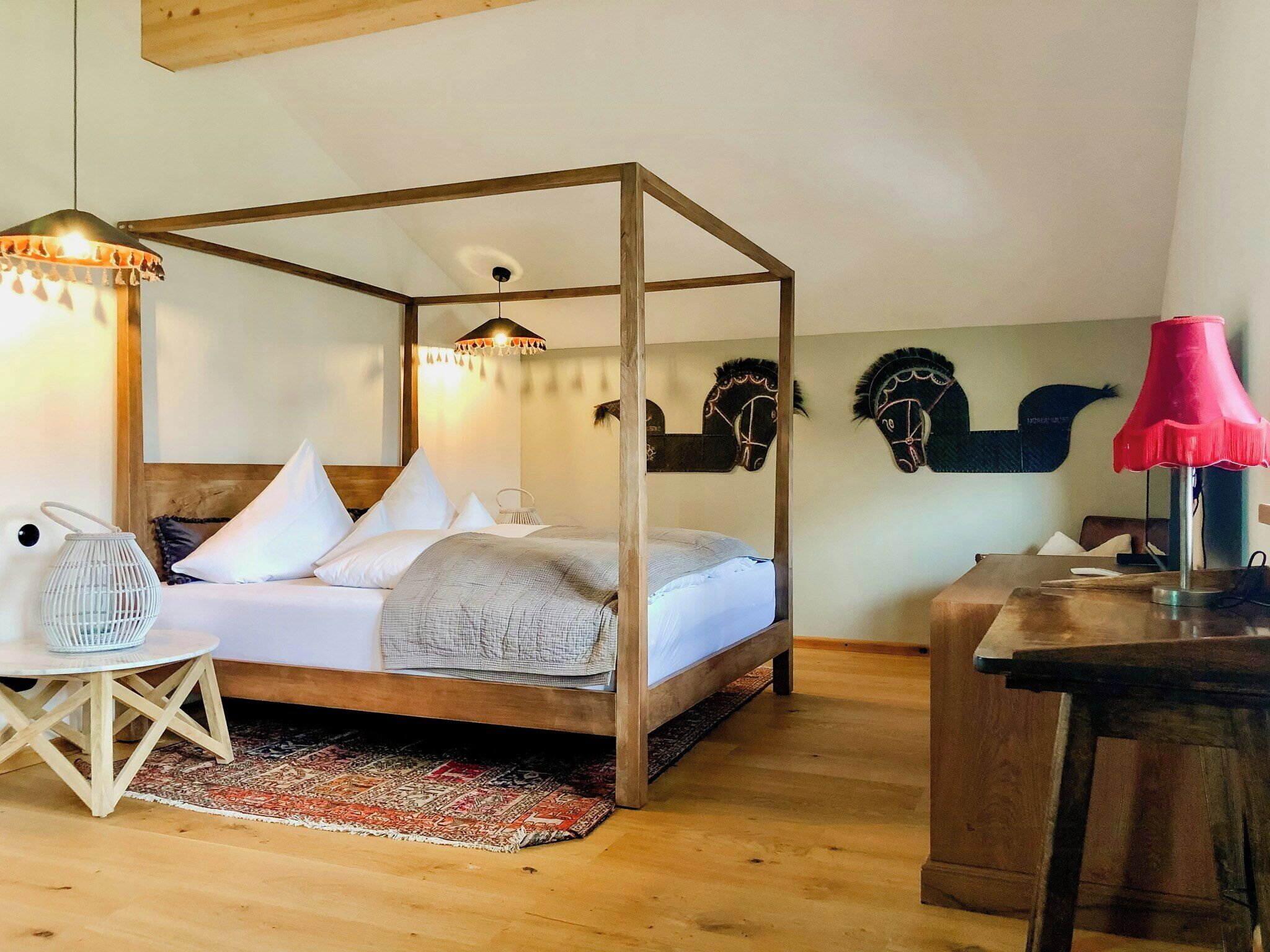 A four-poster bed with white bedding, a wooden floor, a small round table with a white chair, and a red lamp on a wooden desk.