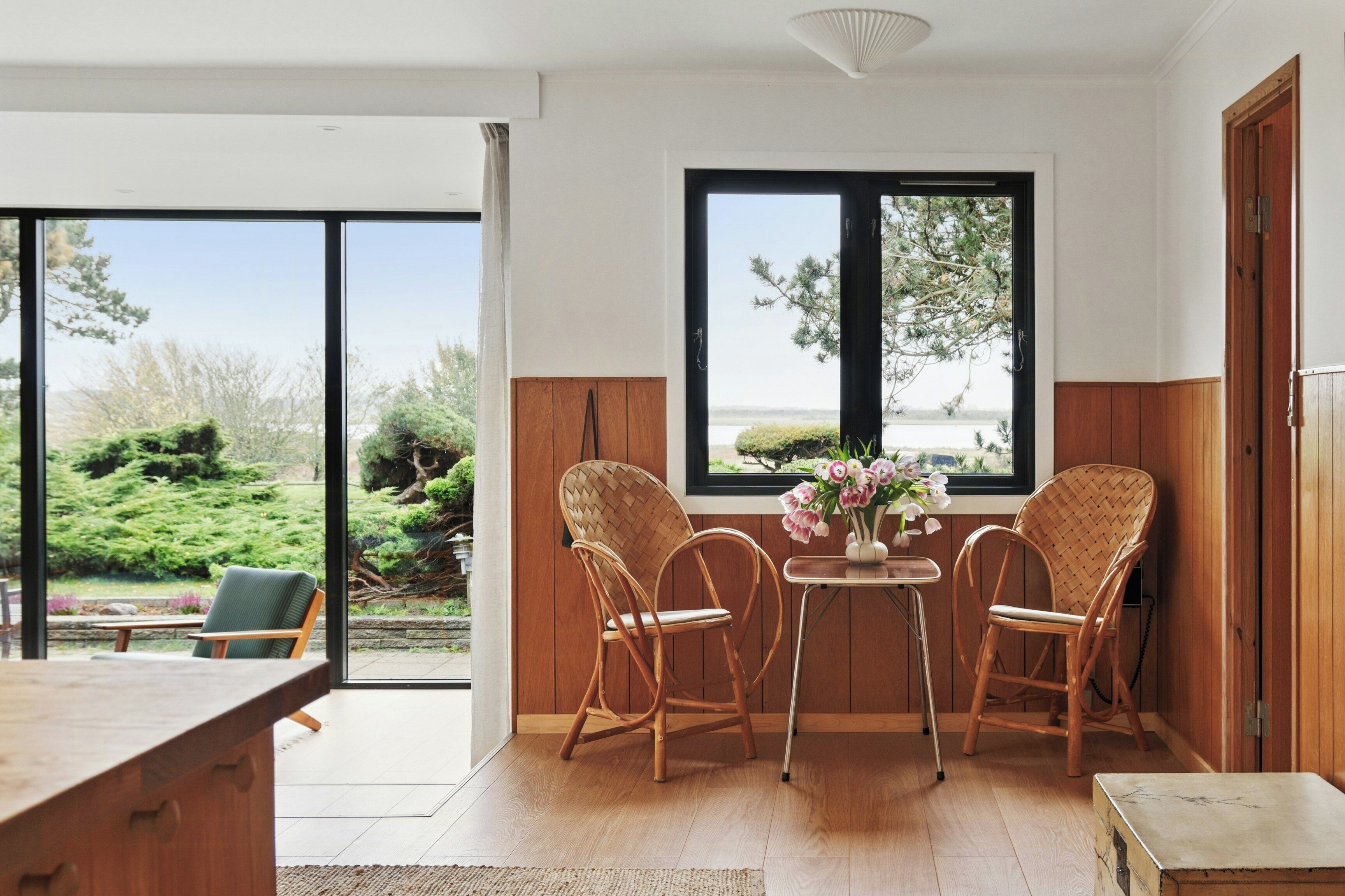 Une pièce avec une grande fenêtre, deux fauteuils en osier, une petite table ornée de fleurs et un bureau en bois.