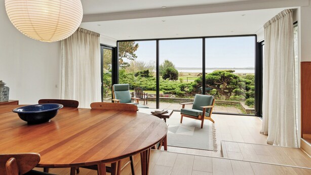 A modern dining area with a wooden table, chairs, and a large window offering a view of the outdoors. - 70s holiday home with views of Vesterlyng (Føllenslev)