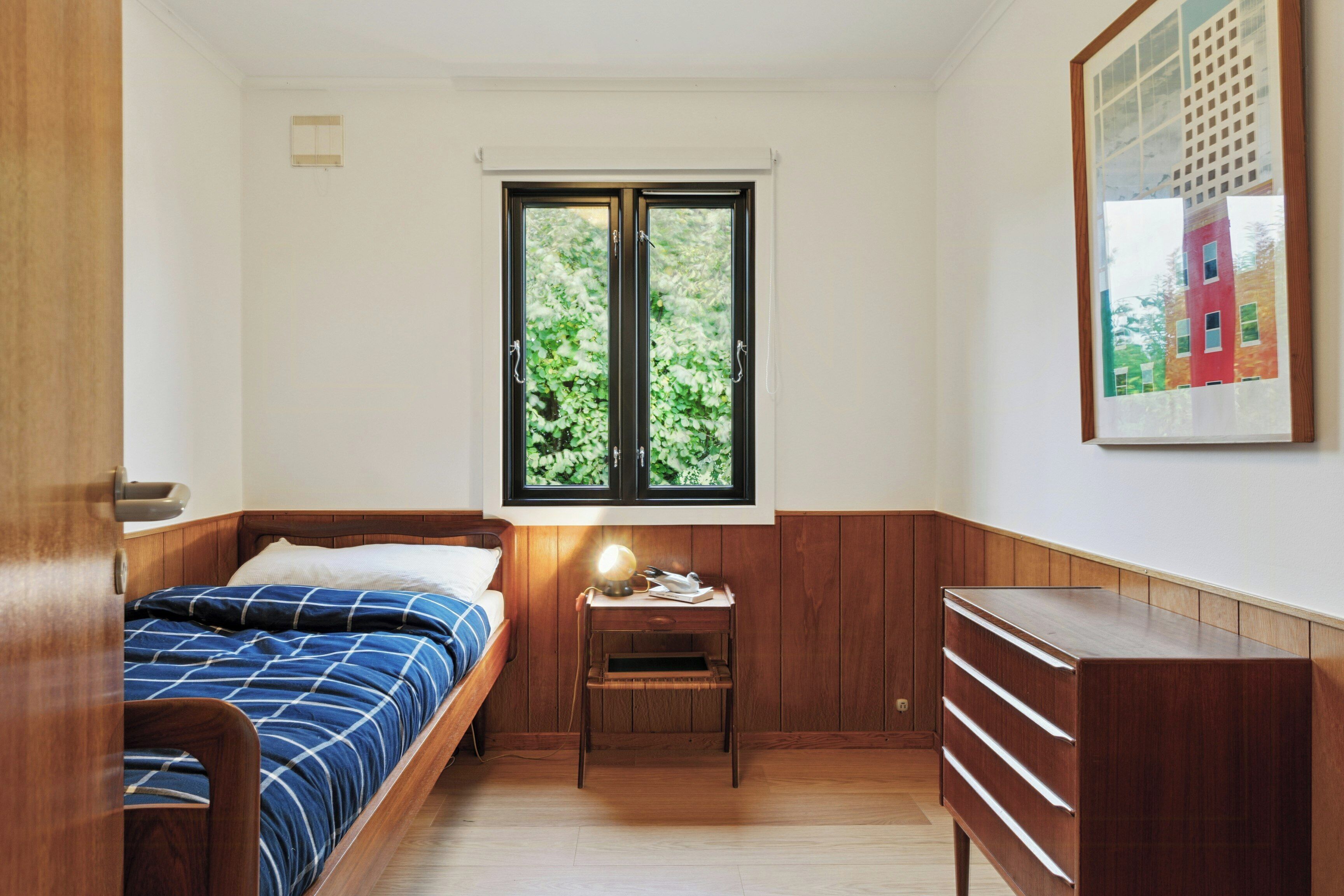 Une chambre à coucher avec un lit, une table de chevet avec une lampe, une commode et une fenêtre donnant sur des arbres.
