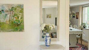 Una habitación con pared blanca, un cuadro, un jarrón con flores, una silla blanca, un mantel estampado y un espejo.