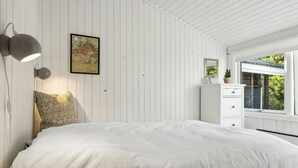 Un dormitorio con cama, mesita de noche, lámpara de pared, cuadro enmarcado, cómoda y ventana con vistas.