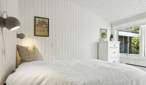 Ein Schlafzimmer mit Bett, Nachttisch, Wandlampe, gerahmtem Bild, Kommode und Fenster mit Blick.