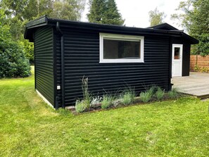 A small, black wooden cabin with a white door and window, surrounded by a well-maintained lawn and a wooden deck. - Summerhouse close to nature and beach (Rørvig)