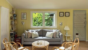 Un salón acogedor con un sofá gris, sillones de mimbre, una mesita redonda y un ventanal con vistas a la vegetación.