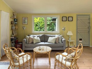 Ein gemütliches Wohnzimmer mit einem grauen Sofa, Korbstühlen, einem kleinen runden Tisch und einem Fenster mit Blick auf Grünflächen.