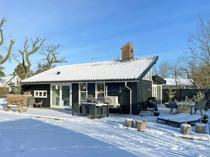 Ein Haus mit schneebedecktem Dach, einem Backsteinkamin und einer überdachten Terrasse mit Gartenmöbeln.