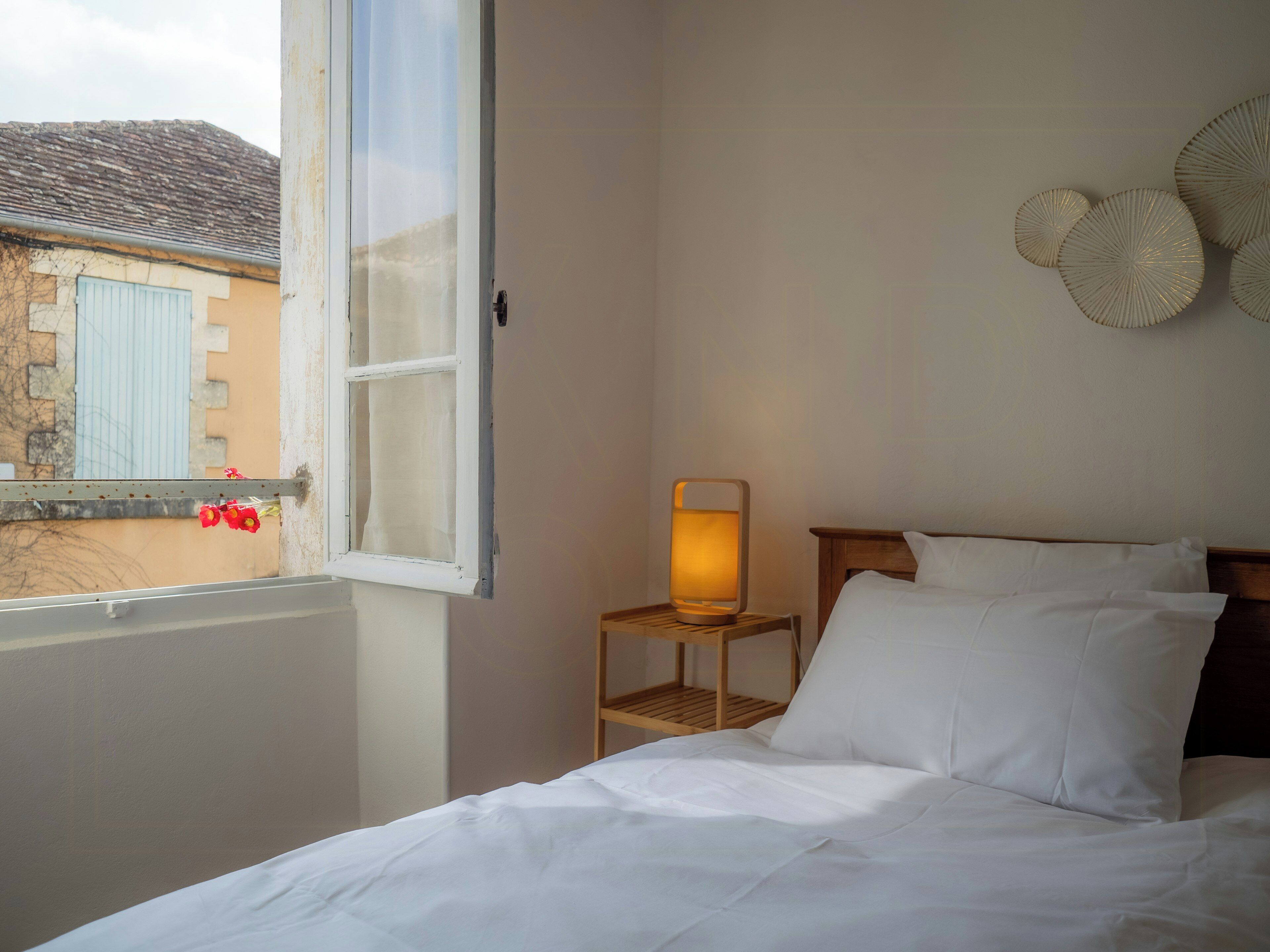Une chambre à coucher avec un lit, une table de chevet avec une lampe, une fenêtre avec des rideaux et un élément de décoration murale.