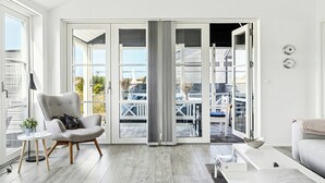 Ett modernt vardagsrum med en grå fåtölj, en vit soffa, ett litet bord med en växt och en stor skjutdörr i glas som leder ut till ett utomhusområde.
