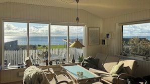 Een gezellige kamer met een groot raam met uitzicht op een mooi panorama, een rieten stoel, een comfortabele bank en een kleine tafel met decoratieve voorwerpen.