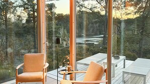 Ein modernes Interieur mit zwei Holzstühlen, einem kleinen Tisch und einer Stehlampe. Große Fenster bieten Blick auf ein bewaldetes Gebiet.