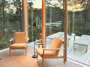 Ein modernes Interieur mit zwei Holzstühlen, einem kleinen Tisch und einer Stehlampe. Große Fenster bieten Blick auf ein bewaldetes Gebiet.