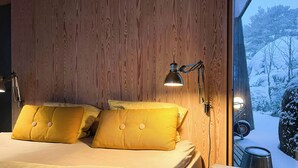 Ein Schlafzimmer mit Holzwandverkleidung, einem Bett mit gelben Kissen, Nachttischlampen und Blick auf eine verschneite Landschaft durch ein Fenster.