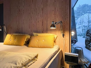 Ein Schlafzimmer mit Holzwandverkleidung, einem Bett mit gelben Kissen, Nachttischlampen und Blick auf eine verschneite Landschaft durch ein Fenster.