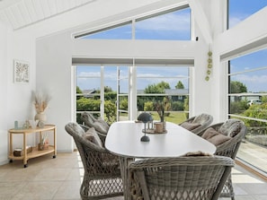 A spacious room with a large dining table surrounded by wicker chairs, large windows offering a view of the outdoors, and a small side table with decorative items.