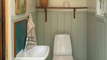 Un baño pequeño con un lavamanos, inodoro y cubo de basura blancos, suelo verde y un estante de madera con una planta y toallas.