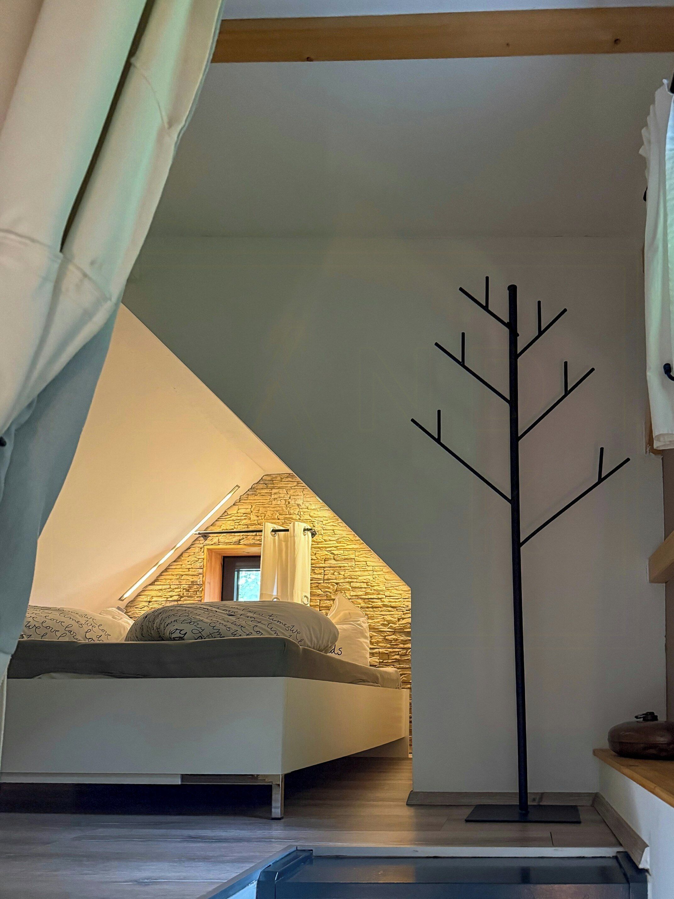 A bedroom with a sloped ceiling, a bed, a coat rack, and a small window.