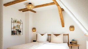 Une chambre à coucher avec un lit, des poutres apparentes, une étagère fixée au mur avec des livres et deux appliques murales.