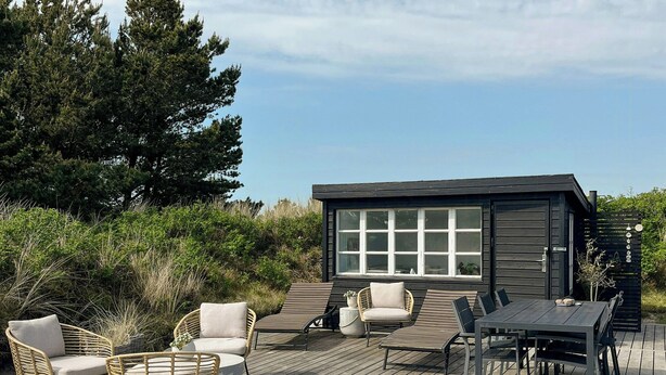 A wooden deck with outdoor furniture, a small building with large windows, and surrounding greenery. - House among dunes and trees in Søndervig (Ringkøbing)