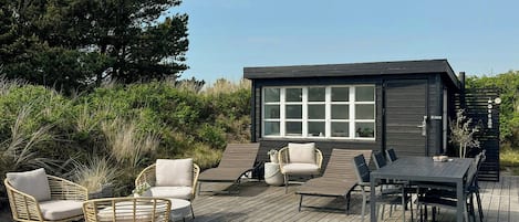 A wooden deck with outdoor furniture, a small building with large windows, and surrounding greenery.
