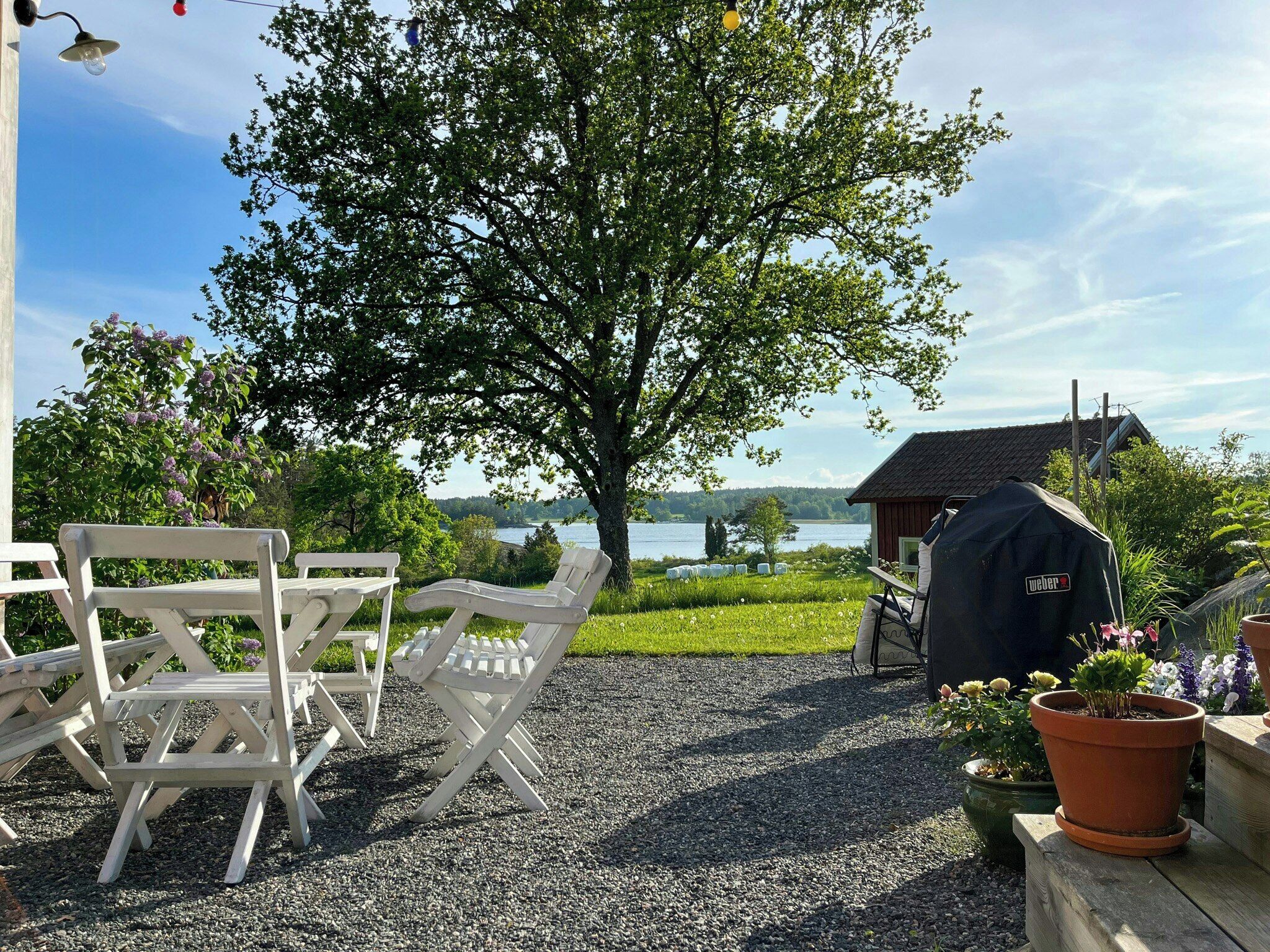 A patio with white chairs, a barbecue grill, and a view of a lake and house.