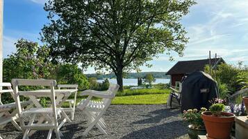 A patio with white chairs, a barbecue grill, and a view of a lake and house.