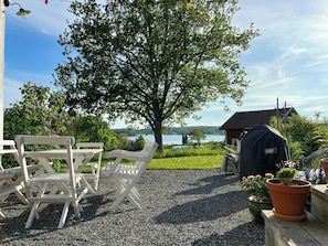 Eine Terrasse mit weißen Stühlen, einem Grill und Blick auf einen See und ein Haus.