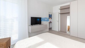 Quarto moderno com cama, televisão, guarda-roupa e janela com cortinas translúcidas.