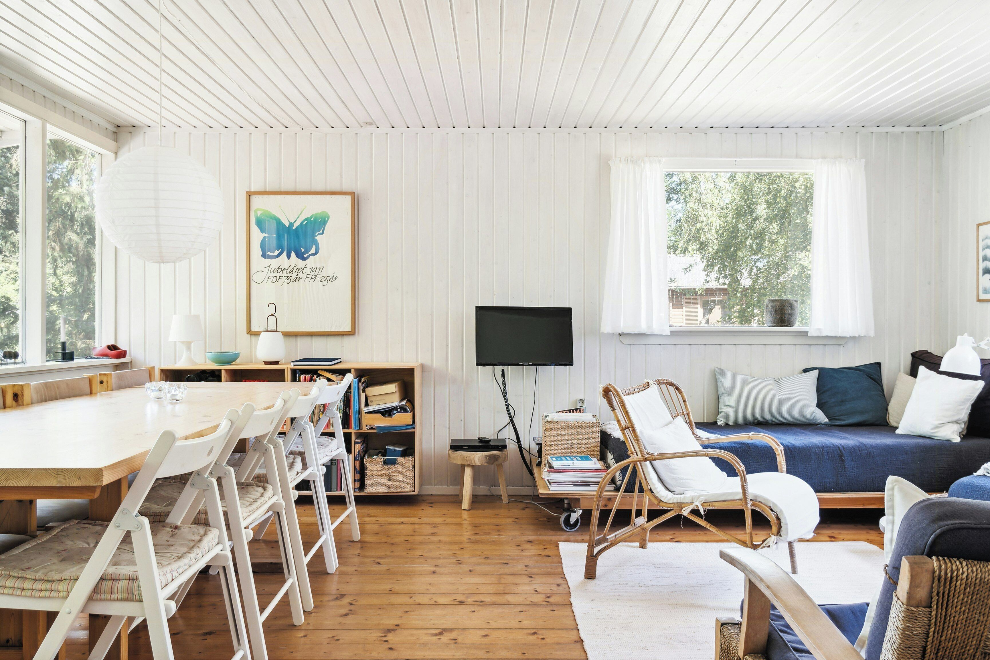 A room with a wooden dining table, white chairs, a sofa, a TV, and a bookshelf.