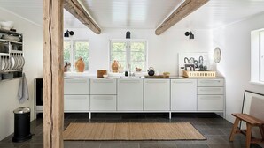 A kitchen with white cabinets, a wooden beam ceiling, a window with a view, and a black trash bin.