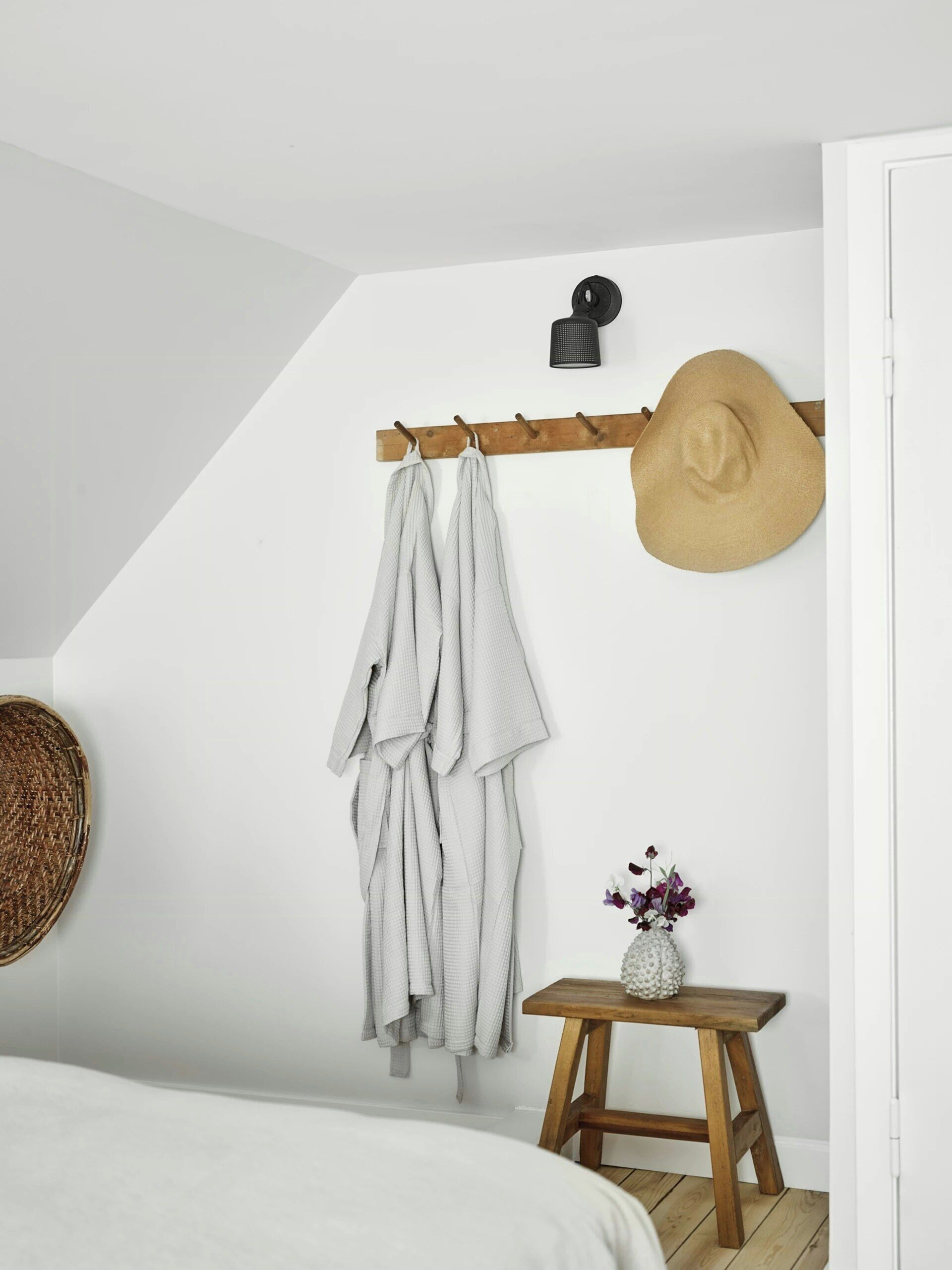 A bedroom with a bed, a wooden stool with a vase, a hat on a coat rack, and a woven basket on the wall.