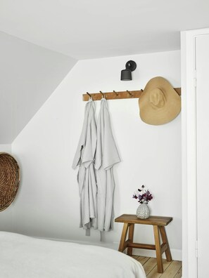 Une chambre avec un lit, un tabouret en bois sur lequel est posé un vase, un chapeau sur un porte-manteau et un panier tressé accroché au mur.
