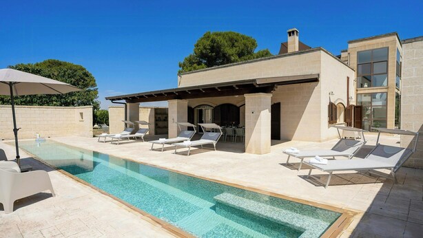 Una piscina con sillones de descanso, una pérgola y un edificio de dos pisos con ventanas amplias.