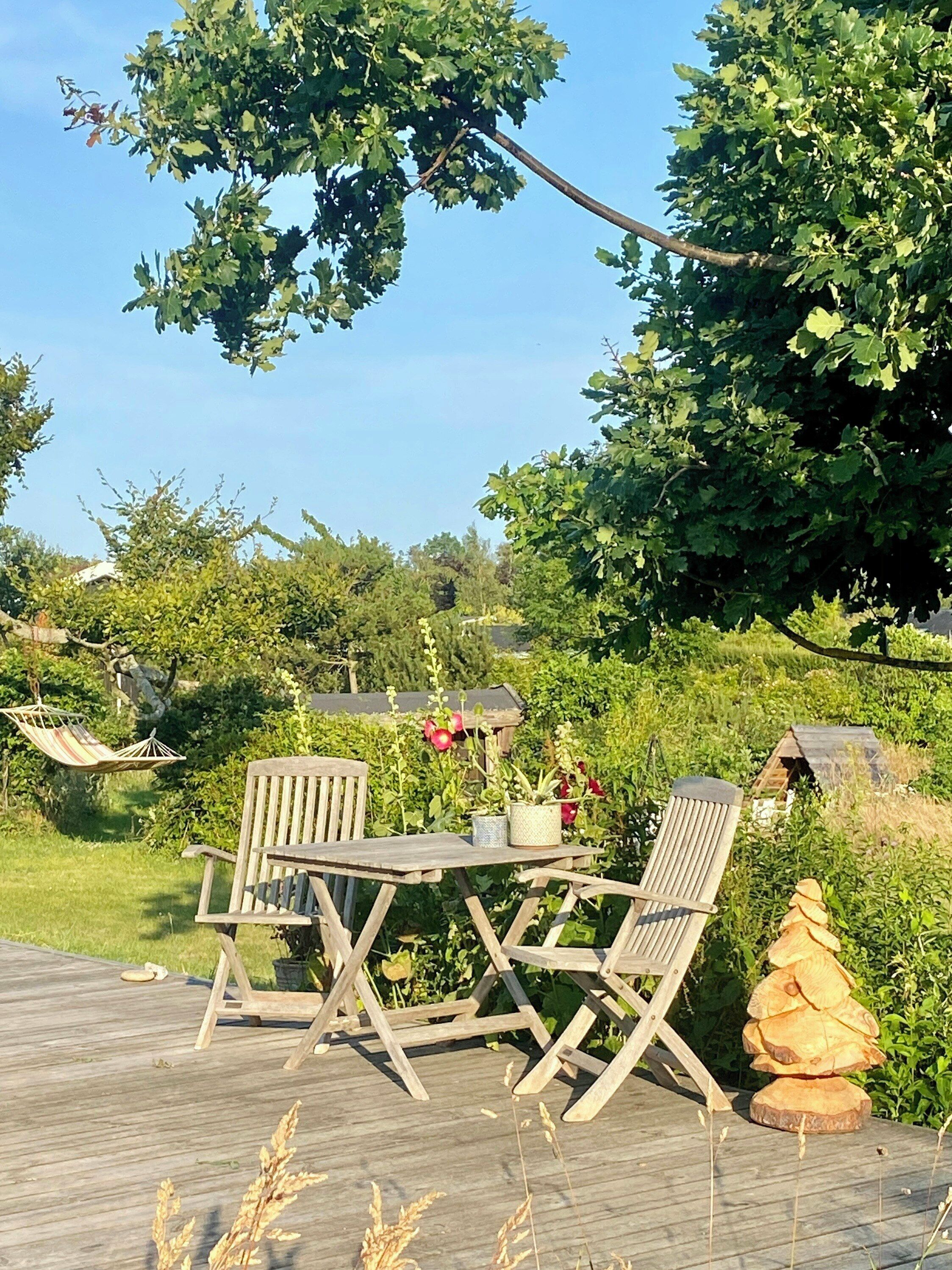 Une terrasse en bois avec une table et des chaises, entourée de verdure et agrémentée d’un hamac.