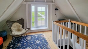 A room with a sloped ceiling, a window with a view of a green landscape, a white crib with a wooden railing, a wicker chair with a fluffy cushion, and a patterned rug on the floor. - Maritime holiday home secluded on the dike (Osterhever)