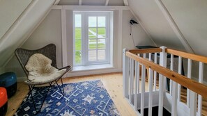 Ein Zimmer mit schrägen Wänden, einem Fenster mit Blick auf eine grüne Landschaft, einem weißen Kinderbett mit Holzgitter, einem Korbstuhl mit einem flauschigen Kissen und einem gemusterten Teppich auf dem Boden.