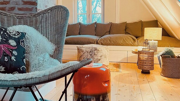 A cozy loft-style room with a high ceiling, a brick wall, a wicker chair, a sofa, a small orange stool, a patterned rug, and a window with a view of trees. - Maritime holiday home secluded on the dike (Osterhever)