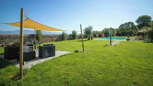 Un patio con césped, piscina, mobiliario exterior y una zona con sombra.