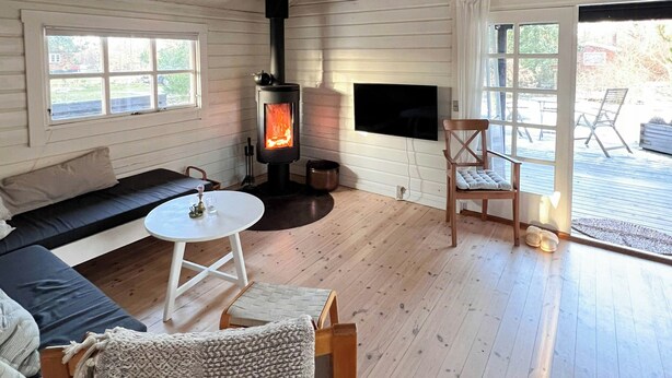 Interior acolhedor de uma cabana, com lareira a lenha, um sofá, uma TV e uma mesa de jantar.