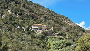 A two-story house with a balcony, surrounded by dense vegetation and rocky terrain. - Secluded villa with sea view in Palombaggia (porto vecchio)