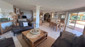 A spacious living area with a sofa, coffee table, and dining table. There is a TV and a kitchen in the background. - Secluded villa with sea view in Palombaggia (porto vecchio)