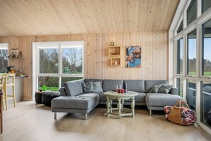 Ein gemütliches Wohnzimmer mit einem grauen Ecksofa, einem runden Couchtisch und einem großen Fenster mit Blick ins Freie.