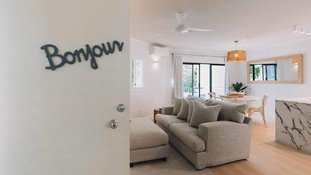 En moderne stue med sofa, spiseplads og et skilt med teksten 'Bonjour' på døren.